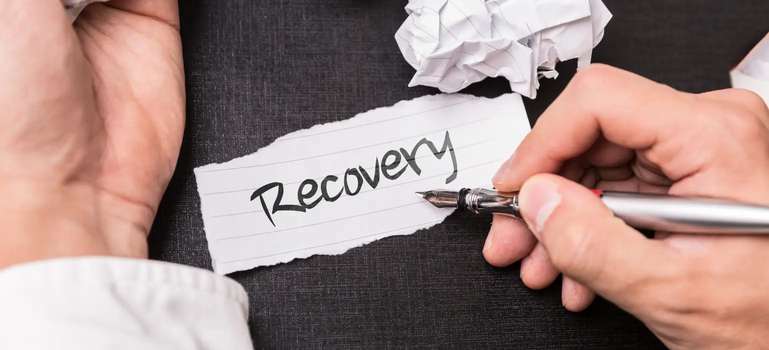 Person writing “Recovery” on paper with Vanderburgh Sober Living wreath icon symbolizing support after treatment