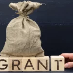 Hand placing a grant block beside a funding bag, representing sober living operator training support in Massachusetts